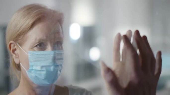 戴着安全面具的患病老年妇女在医院通过防护玻璃墙与家人访客交谈。