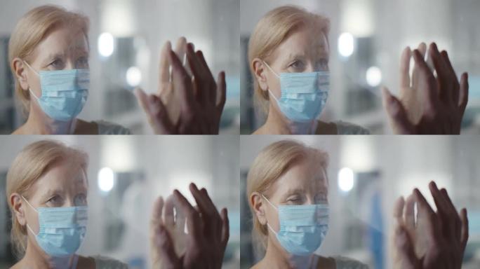 戴着安全面具的患病老年妇女在医院通过防护玻璃墙与家人访客交谈。