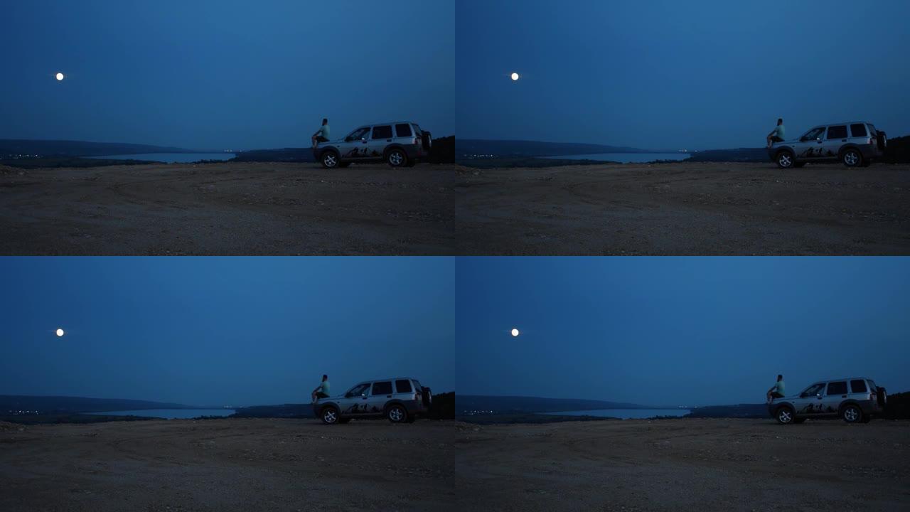 一个人独自坐在河边的汽车引擎盖上，在夜空下凝视着远方
