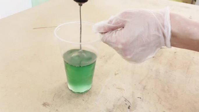 戴手套的手在塑料杯中搅拌绿色苛性碱液体，用于粘合碳的工业液体，该混合物会干扰生产混合器