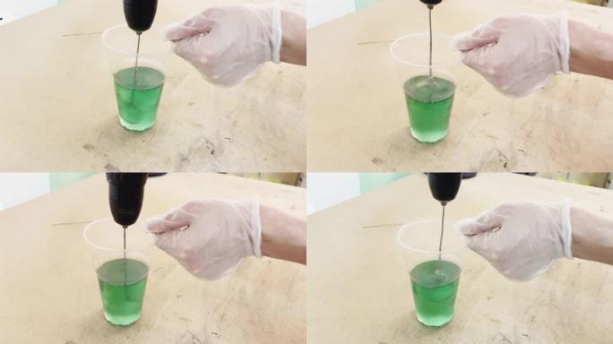戴手套的手在塑料杯中搅拌绿色苛性碱液体，用于粘合碳的工业液体，该混合物会干扰生产混合器