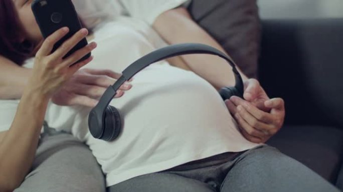 快乐的亚洲丈夫和怀孕的妻子一起度过时光，为未出生的婴儿打开音乐，并躺在家里客厅的沙发上使用智能手机。