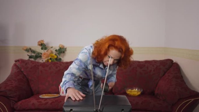 恼怒的复古女人用天线坐在沙发上看电影吃垃圾食品打老式电视。20世纪80年代20世纪70年代周末在家休