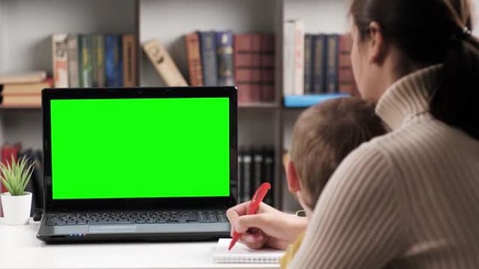 女人和婴儿坐在桌子旁，看着笔记本电脑的绿屏，母亲正在谈论在线视频连接，并在笔记本上做笔记。色度键电脑