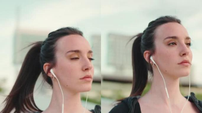 垂直视频: 戴着黑色运动连帽衫的雀斑和长马尾辫的年轻女子戴着有线耳机听音乐并沿着桥走