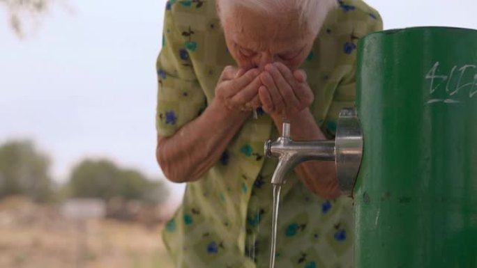 高级女退休人员在户外的水龙头上站着手掌喝水。前视图老高加索皱纹女人在夏日享受清爽的液体。