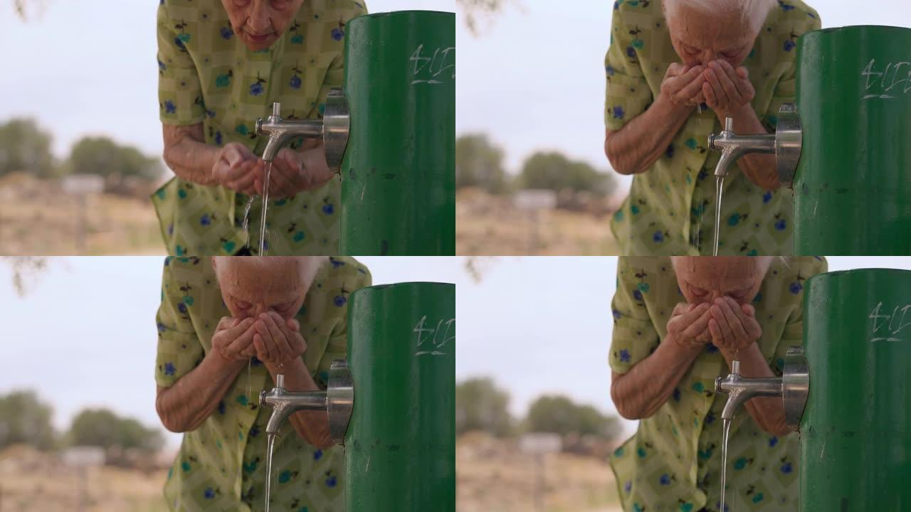 高级女退休人员在户外的水龙头上站着手掌喝水。前视图老高加索皱纹女人在夏日享受清爽的液体。