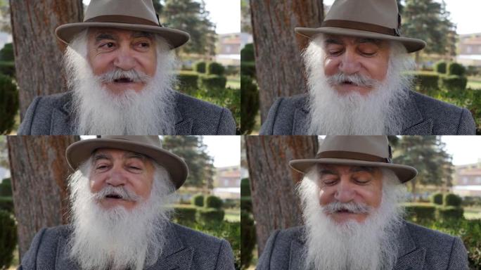 一位老人的肖像，留着蓬松的灰色胡须，戴着一顶好帽子，微笑着坐在公园的长椅上。善良可爱的祖父看起来像童