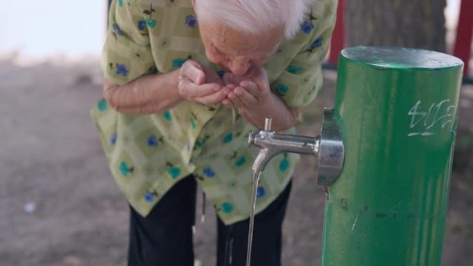 炎热阳光明媚的夏日，白发老妇从室外水龙头喝水。高级高加索退休人员喜欢在南部国家旅游。