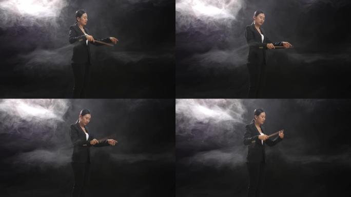 亚洲指挥家女子手持指挥棒，在迷雾笼罩的黑色工作室中迅速展示手势的侧视图