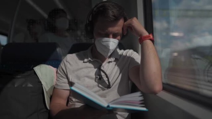 白人男子戴着口罩和耳机坐在火车上，坐在窗户旁看书。男性阅读书籍，并在火车上的耳机上听音乐。主题穿越德