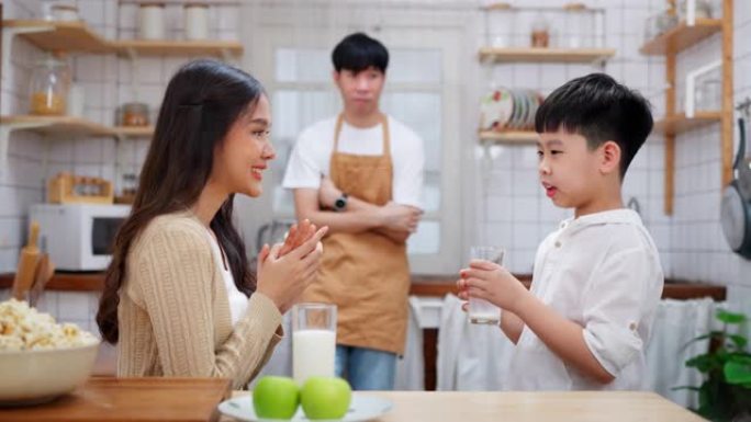 年轻的亚洲男孩在现代厨房里和母亲一起喝牛奶，微笑着，母亲拍手。父亲站在后面，看着家人。享受早餐的幸福