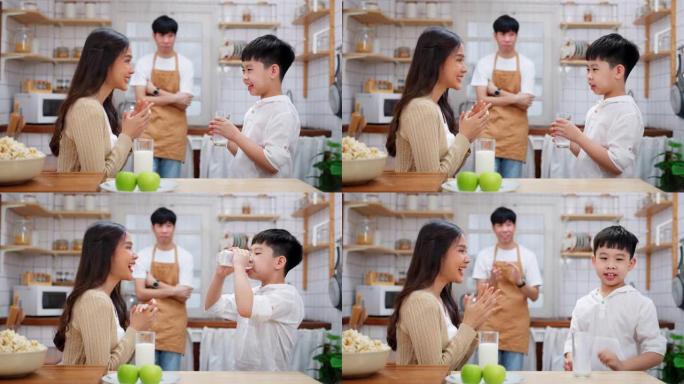年轻的亚洲男孩在现代厨房里和母亲一起喝牛奶，微笑着，母亲拍手。父亲站在后面，看着家人。享受早餐的幸福