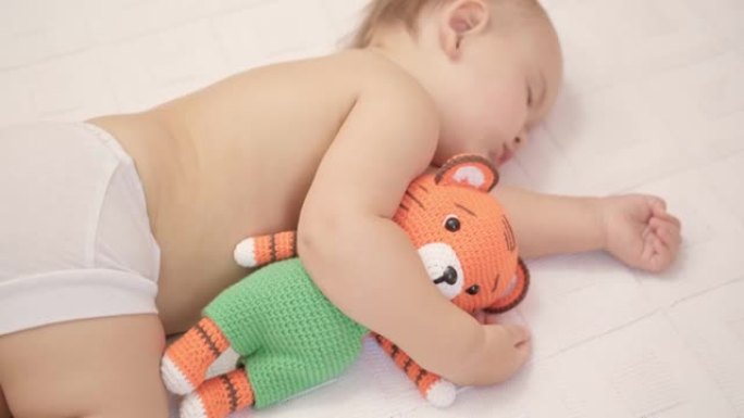 12-17个月大的幼儿睡在床上，抱着一个针织老虎玩具。顶视图