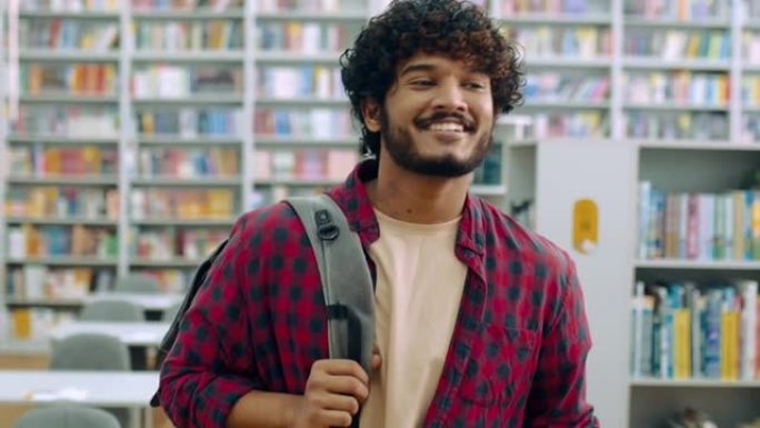 英俊的骄傲的印度或阿拉伯大学男生，穿着休闲装，背着背包和笔记本电脑，在书架的背景下穿过图书馆，看着相