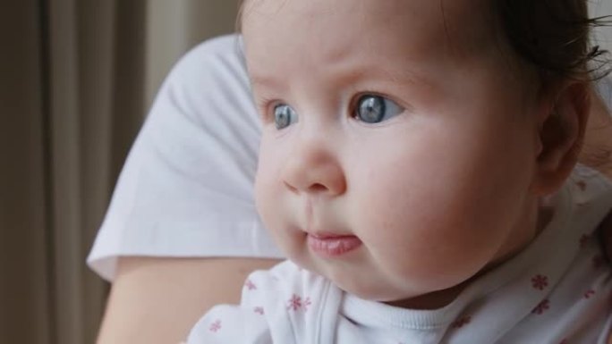 可爱的可爱的婴儿新生儿，蓝眼睛在母亲的怀抱中，富有表现力地看着窗外，动了动她的嘴唇。生命的第三个月特