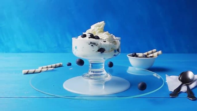 蓝莓落入装有冰淇淋的玻璃杯中，在蓝色背景下的木制桌子上的托盘上旋转。具有速度斜坡效果的4k慢动作视频