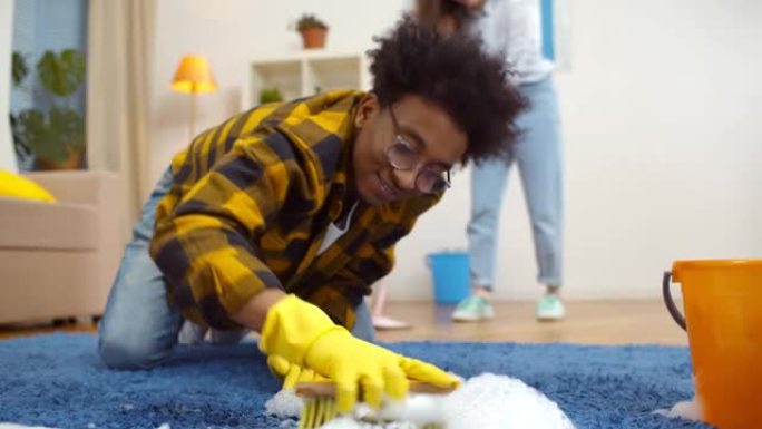 幸福多样的已婚夫妇一起打扫公寓。非裔美国人用洗涤剂和刷子清洁地毯，而女友在背景上拖地