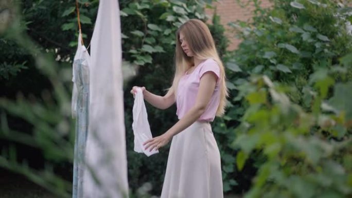 侧视图年轻女子在户外慢动作中挤衣服，将白布挂在绳子上。高加索迷人家庭主妇的肖像在花园里摆姿势看着相机