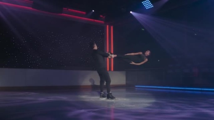 花样滑冰运动员对在聚光灯下进行固定旋转提升。男人抱着女人的两个脚踝，双臂交叉，旋转，她盘旋在冰上，双