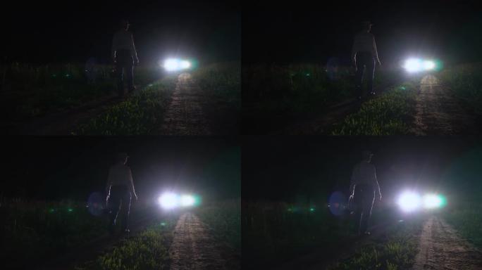 一个拿着篮子和手电筒的男人晚上在田野土路上的汽车前灯下行走。一个男人的剪影在黑暗的午夜在田野的草地上
