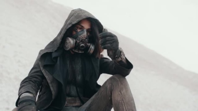 世界末日后的女性流浪者，戴着垃圾头罩，戴着防毒面具，坐在沙漠里，手倒沙子。蒸汽朋克朋克士兵。死亡世界