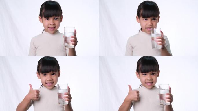 可爱的亚洲小女孩拿着一杯水，在工作室的白色背景上竖起大拇指。儿童良好的健康习惯。医疗保健概念