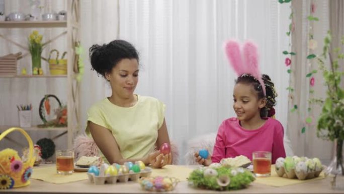 妈妈和女儿玩得开心，打破复活节彩蛋。非裔美国妇女和小女孩坐在家里装饰精美的房间里的桌子旁。复活节快乐