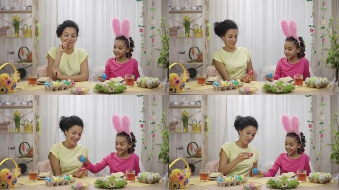妈妈和女儿玩得开心，打破复活节彩蛋。非裔美国妇女和小女孩坐在家里装饰精美的房间里的桌子旁。复活节快乐