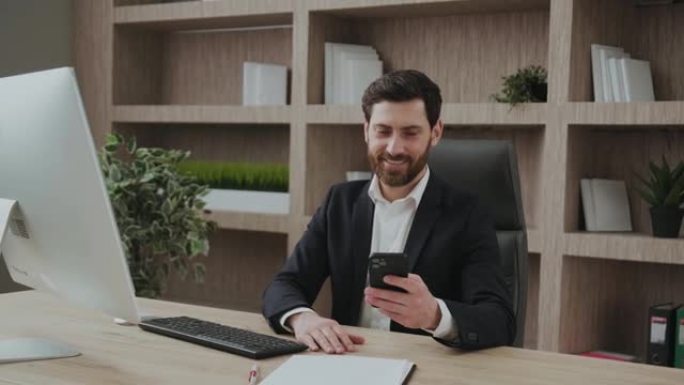 穿着正式西装的成功英俊的执行经理坐在办公桌前看着智能手机，一边微笑着阅读信息或好消息