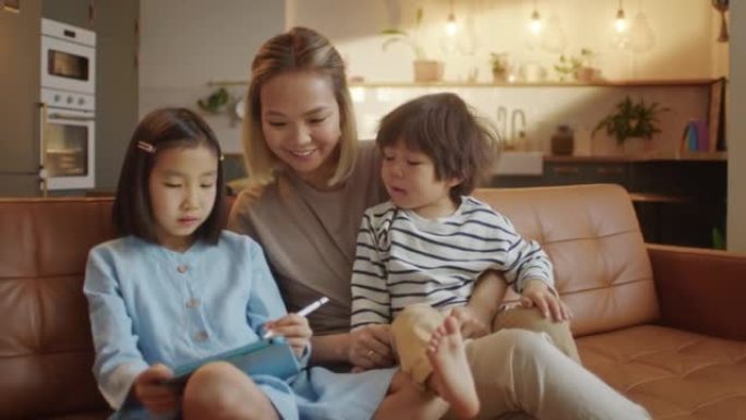 充满爱心的亚洲妈妈和小女儿和儿子一起坐在家里的沙发上，平板电脑设备用铅笔绘制，使用教育应用程序轻松有