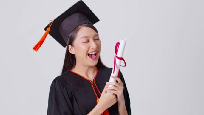美丽迷人的亚洲女人毕业于帽子和礼服微笑，手里拿着认证的感觉如此自豪和幸福，孤立在白色背景上，教育成功
