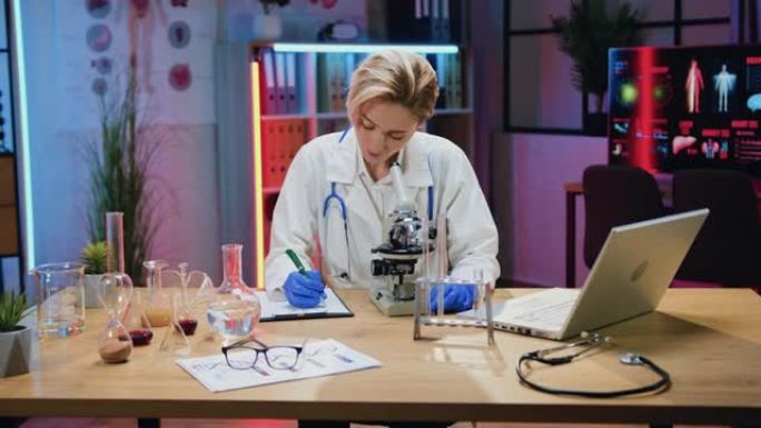 有吸引力的自信的观察力熟练的金发女科学家生物学家，穿着实验服和手套，在显微镜下观察样品，并在论文报告