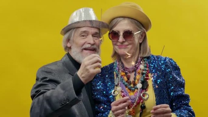 年老的男人和女人。黄色背景上时尚的老年妇女。她戴着帽子和眼镜。戴着帽子胡须的时尚男人。他们手里有烟火