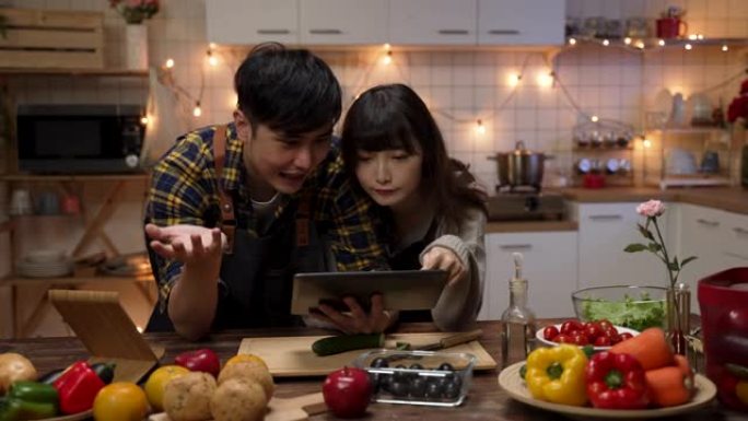 快乐的亚洲情侣在家里的厨房里一起计划和决定情人节晚餐食谱时，使用触摸板