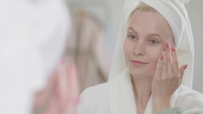 迷人的年轻金发女子的肖像，穿着浴袍，自然化妆，在皮肤上涂上奶油，看着镜子。皮肤护理水疗程序概念的广告