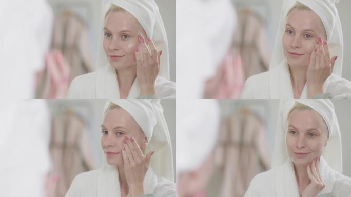 迷人的年轻金发女子的肖像，穿着浴袍，自然化妆，在皮肤上涂上奶油，看着镜子。皮肤护理水疗程序概念的广告