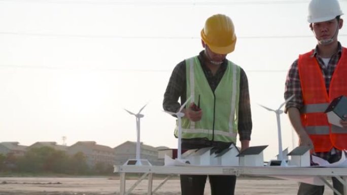 两位电气工程师讨论了建筑工地项目中的家用太阳能电池农场和风力涡轮机模型以及日落时的高压电力线塔。团队