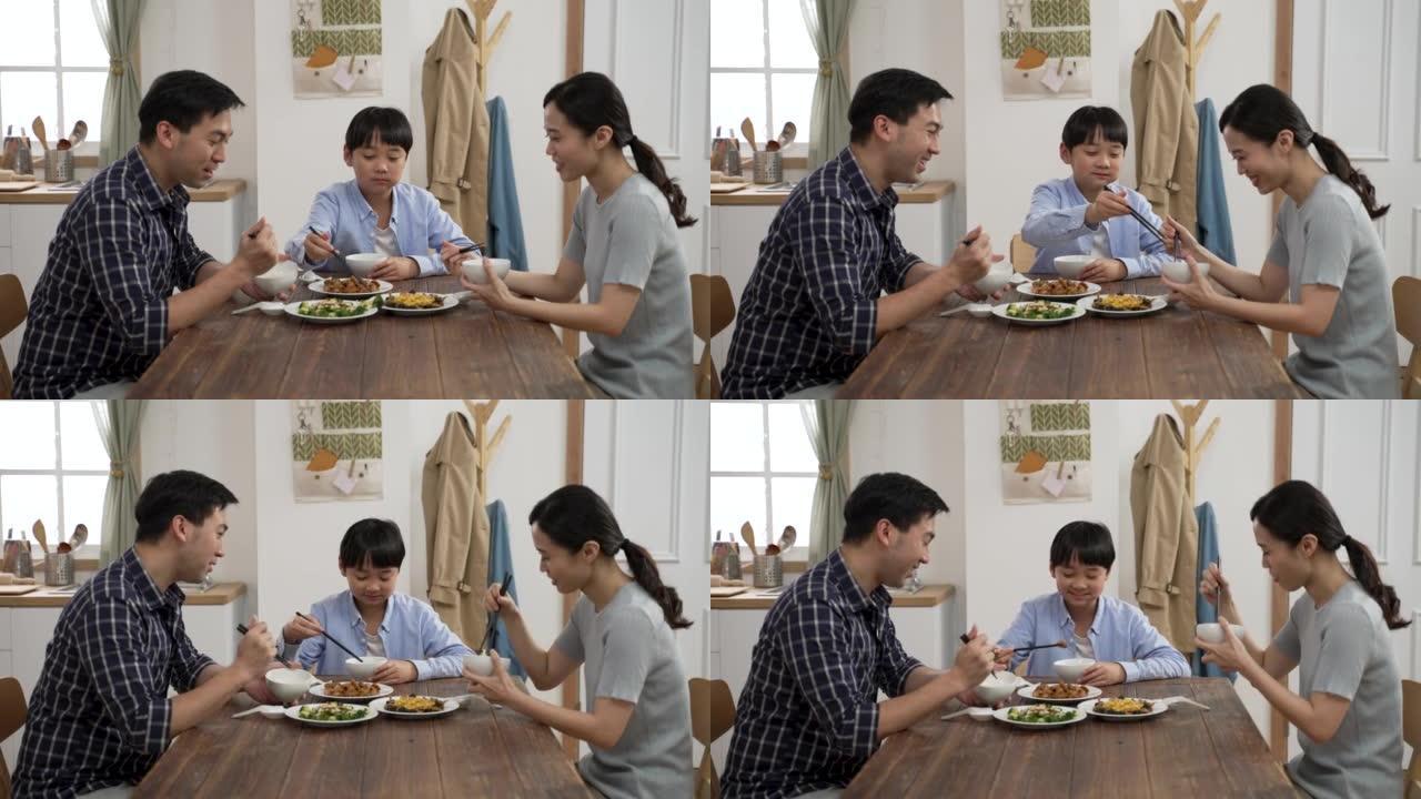 开朗的亚洲爸爸和妈妈在明亮的家庭室内与他们的小儿子在餐桌上享受美味的食物时，互相微笑和点头