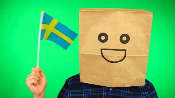 头上戴着纸袋的男子的肖像，在绿色的背景下挥舞着瑞典国旗，面带微笑。