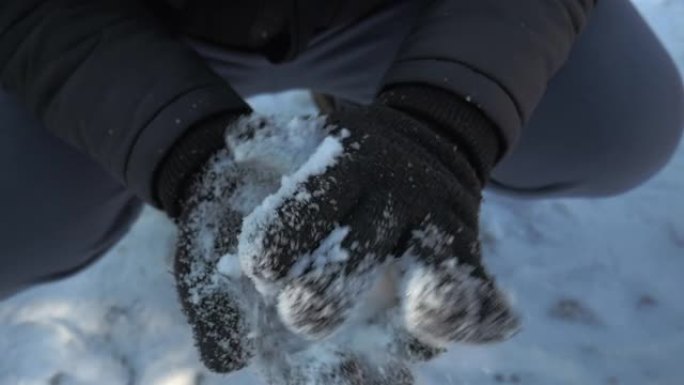 近距离拍摄一个制作雪球的人的手。该人戴着黑色羊毛手套，黑色夹克和蓝色运动服