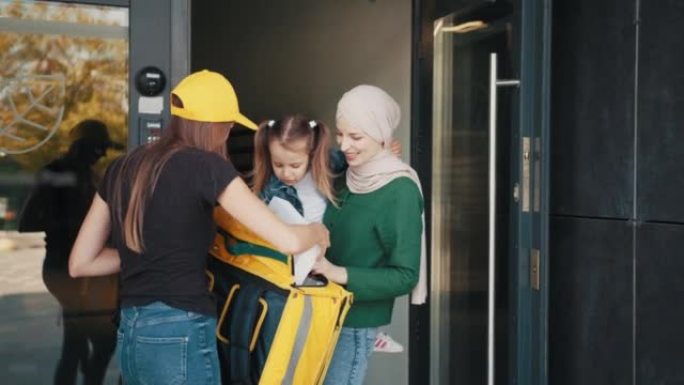 一名戴着头巾的积极的穆斯林妇女带着孩子接受了一个戴着黄色帽子和保暖背包的快递女孩的在线订单。交付概念