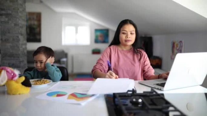 在同一张桌子上坐着一个正在吃早餐的小男孩，他的泰国母亲在家工作