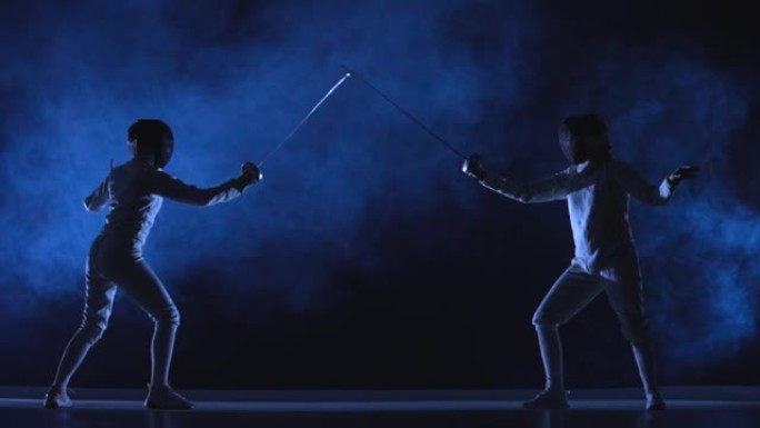 两名女运动员在击剑比赛中进行花剑训练。穿着白色制服和击剑面具的年轻剑客在黑暗的工作室背景下与烟雾和蓝