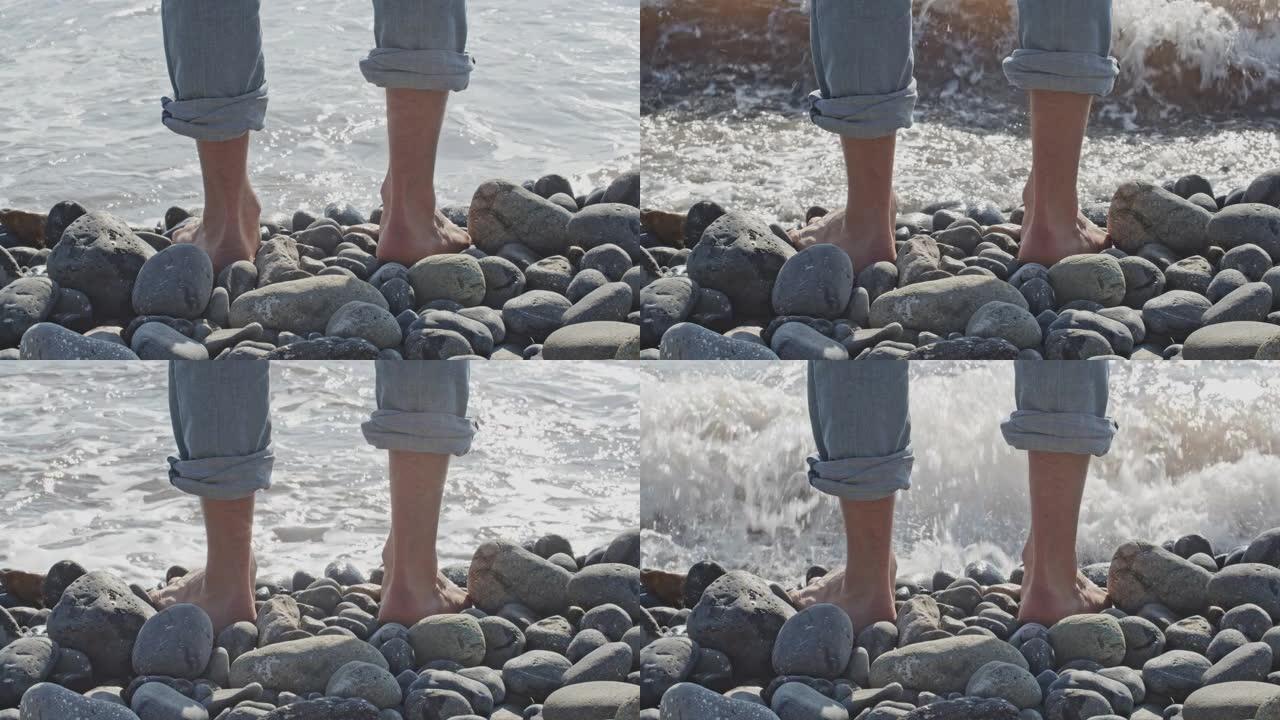 一个人接近海浪。在冲浪和海滩的背景下，穿着打气牛仔裤的男人的腿。后视图。冬天沿海的内向者