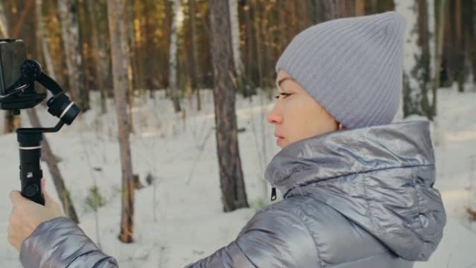 冬季手持薄膜三轴万向节稳定装置的女人为智能手机。摄像师操作员拍摄照片视频