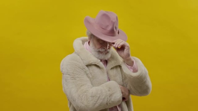 穿着黄色背景粉色衣服的时尚老人。他摸了摸帽子。他的手放在胸前。