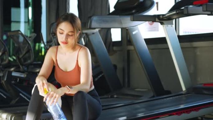 一名亚洲妇女在健身房休息，在健身时间用跑步机跑步后，用毛巾擦干脸，喝矿泉水