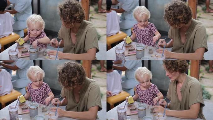 可爱的金发蹒跚学步的孩子和年轻的母亲在艺术工作室学习陶器，一起享受家庭时光。儿童在创造力方面的发展和
