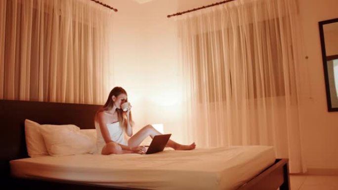 漂亮的白人妇女在舒适的床上休息时喝热茶，在无线笔记本电脑上打字。舒适酒店房间的夜光。生活方式概念。疲